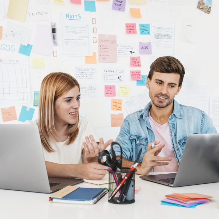 smiling-male-female-sitting-desk-talking-looking-laptop